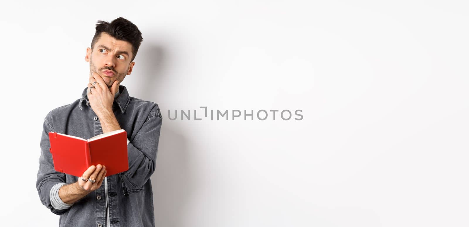 Thoughtful young man plan his schedule, looking aside pensive, holding opened planner or journal, standing on white background.
