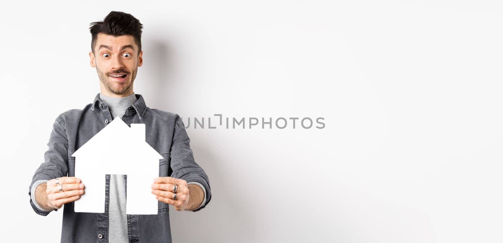 Real estate and insurance concept. Man look with excitement at paper house cutout, buying home, standing on white background.