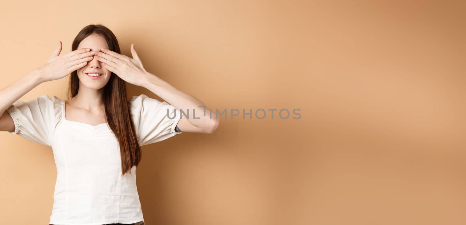 Happy romantic girl covering eyes and waiting for surprise, smiling while anticipating gift, standing on beige background by Benzoix