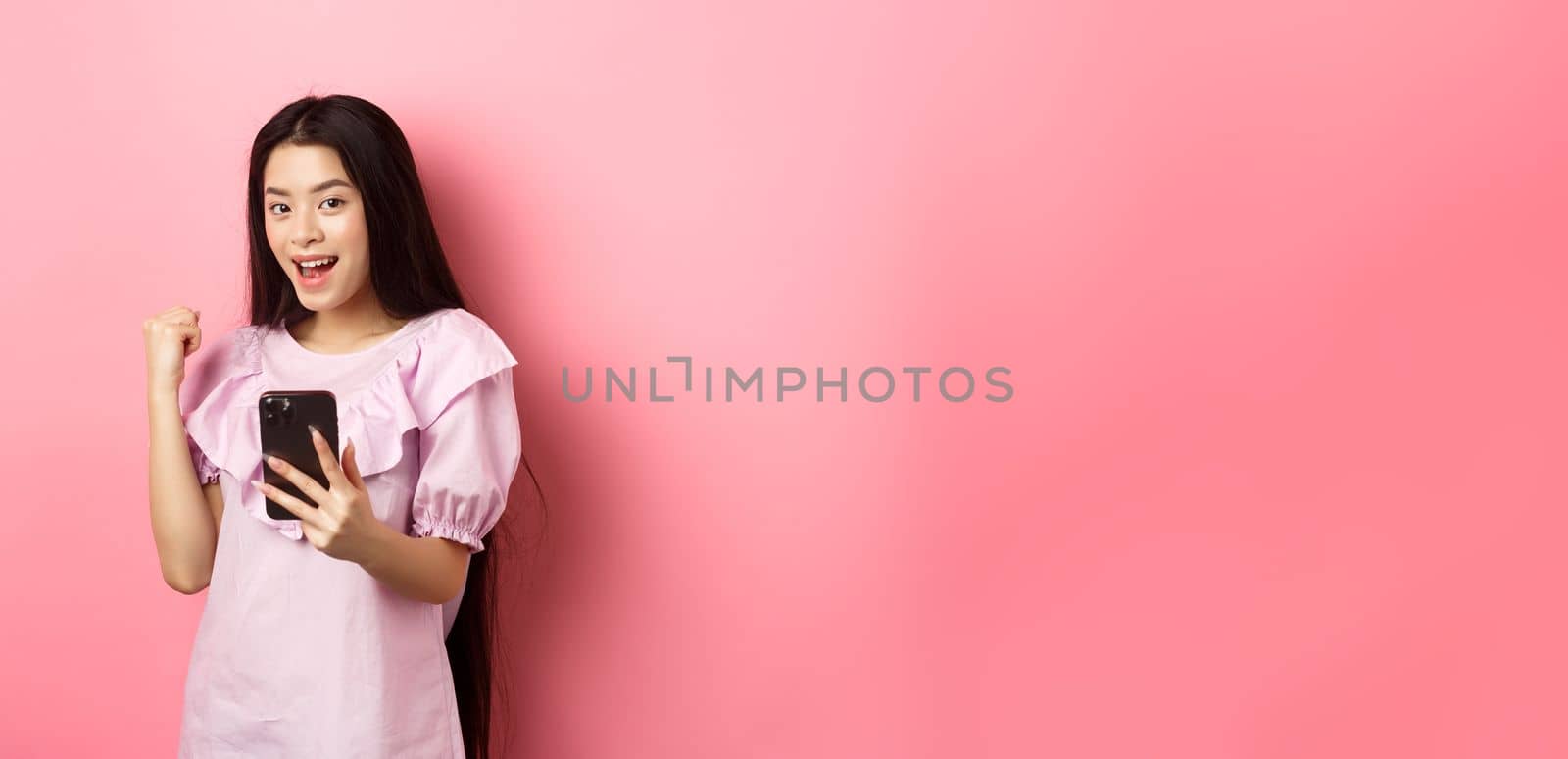 Online shopping. Satisfied asian girl winning on mobile phone, say yes and make fist pump, holding smartphone, standing against pink background by Benzoix