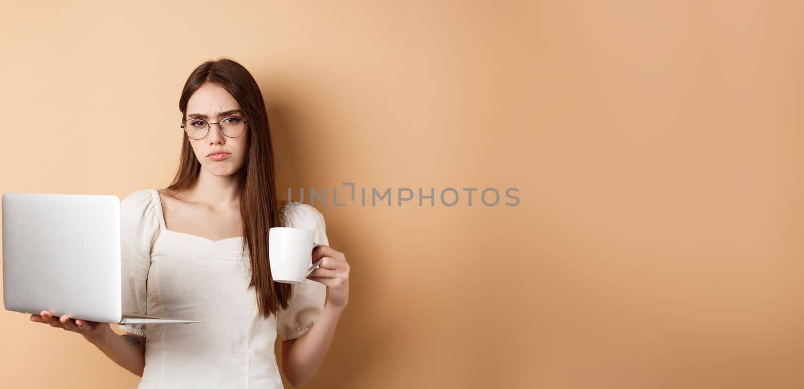 Tired and reluctant girl hates working, drinking morning coffee and using laptop, frowning upset, standing on beige background.