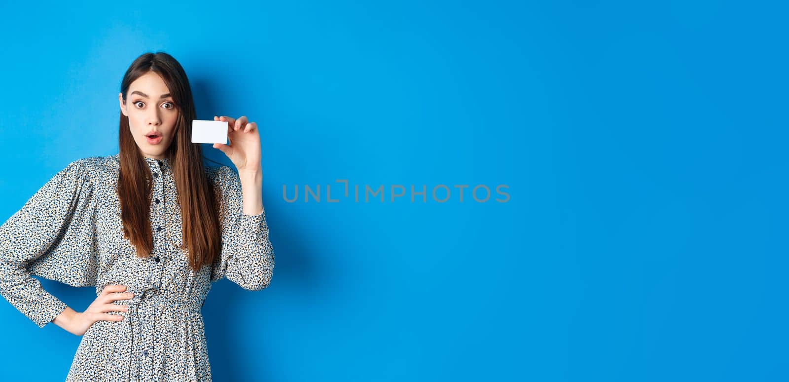 Shopping. Excited girl showing her plastic credit card and look at discounts, say wow amazed, standing on blue background by Benzoix