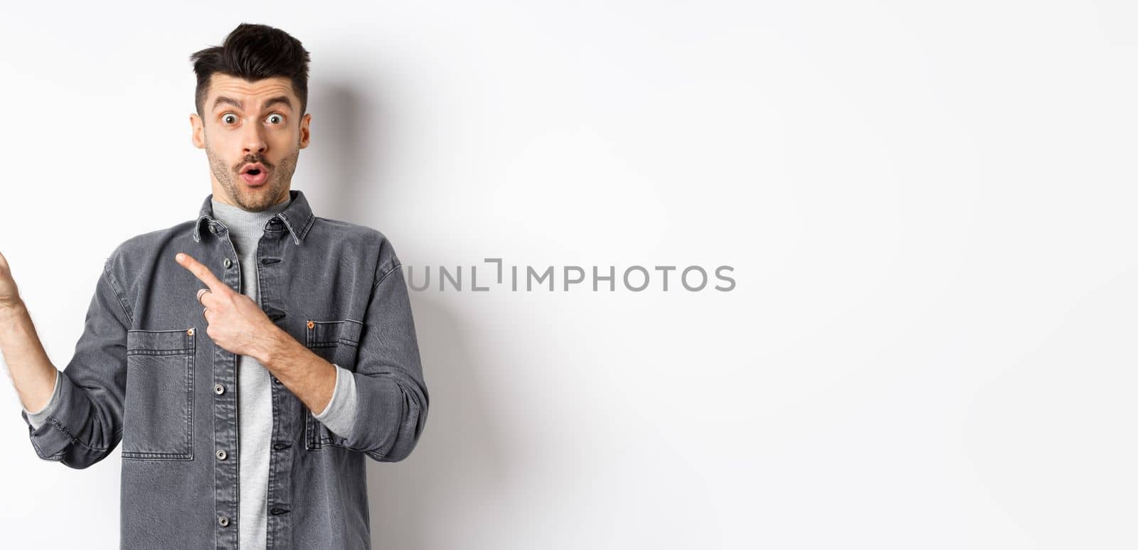 Excited man pointing left at logo and look with disbelief, saying wow at camera, checking out cool promo discount, standing on white background by Benzoix