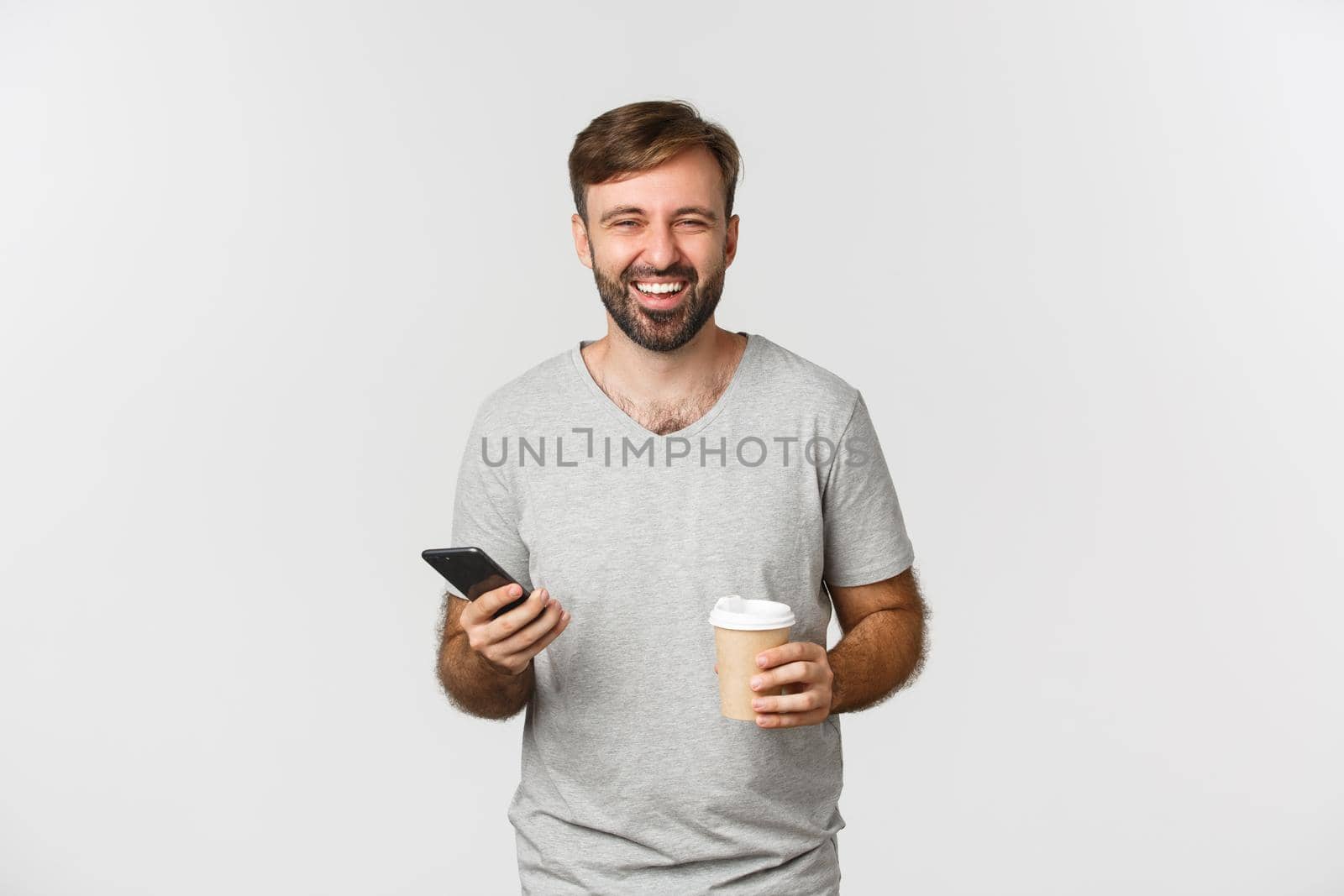 Portrait of carefree attractive man, drinking coffee and using mobile phone, laughing and smiling, standing over white background.