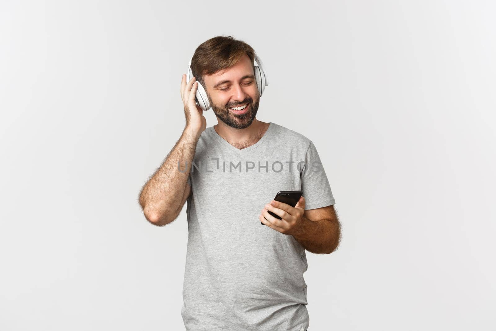 Image of handsome man in casual outfit, listening music in wireless headphones, holding mobile phone, smiling pleased, standing over white background by Benzoix