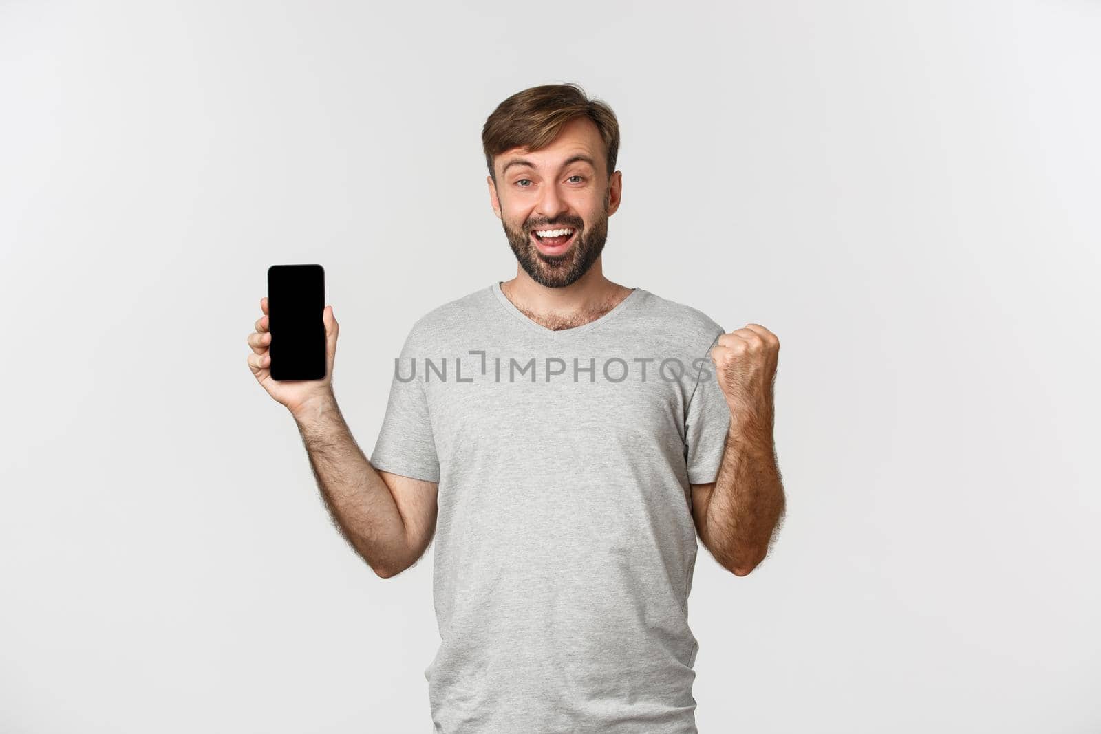 Portrait of excited man with beard, achieve goal in app, showing mobile phone screen and rejoicing, winning something, standing over white background.