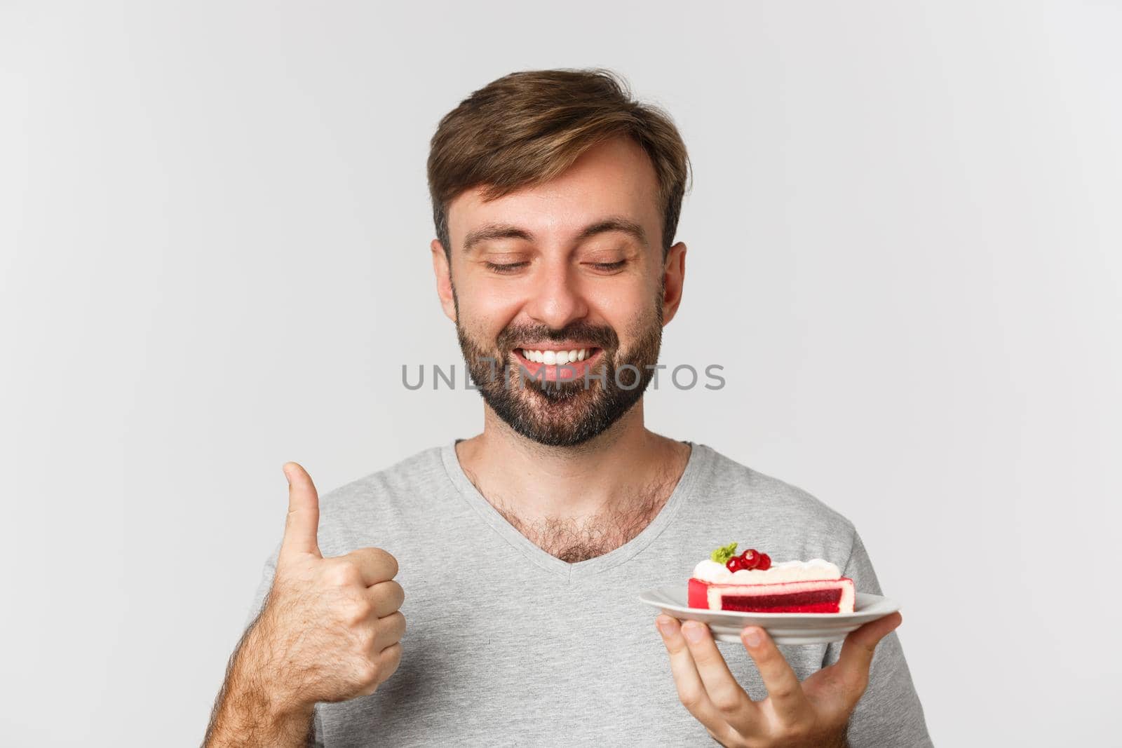 Close-up of cheerful caucasian guy with beard, wearing grey t-shirt, holding cake and showing thumbs-up, like dessert, standing over white background.