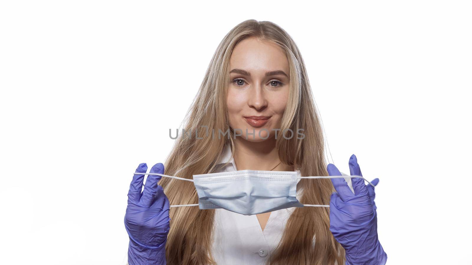 About to put on medical face mask young nurse with long straight hair looking at camera. Caucasian woman wearing white medical uniform isolated on white background.