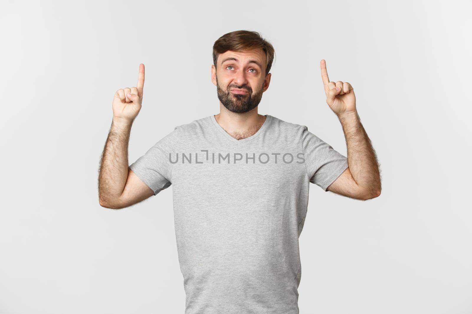 Skeptical and unimpressed adult man with beard, looking up and pouting displeased, standing over white background in gray t-shirt.