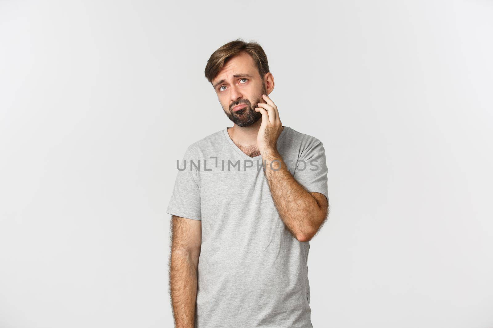 Portrait of indecisive sad man in gray t-shirt, scratching neck and looking up puzzled, standing over white background.