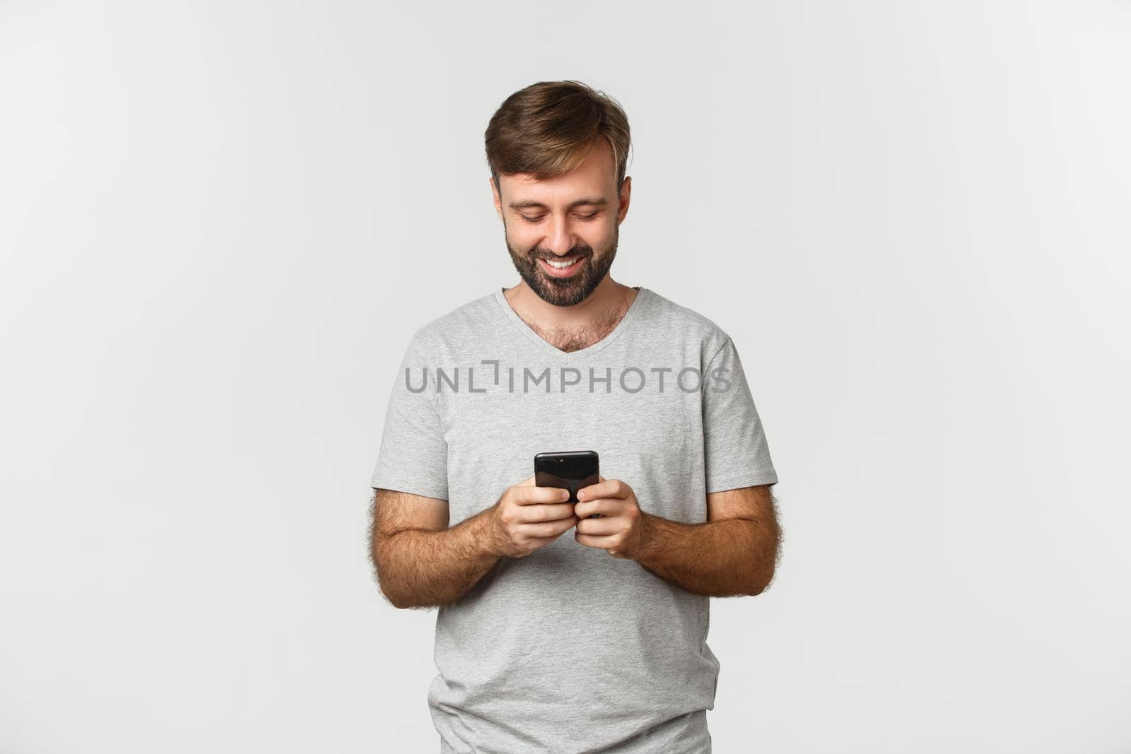 Portrait of handsome modern guy with beard, messaging and looking at smartphone screen, standing over white background.