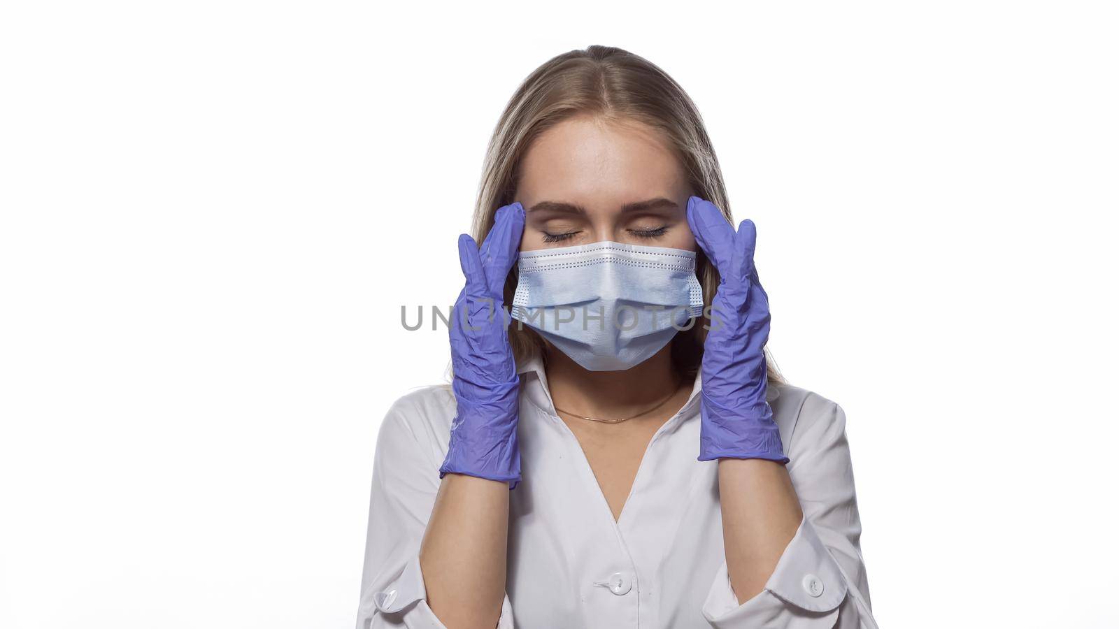 Suffering headache nurse in a medical mask and disposable gloves at work, massaging the temporal lobes of the head, with eyes shut. Isolated on white background. Medicine concept.