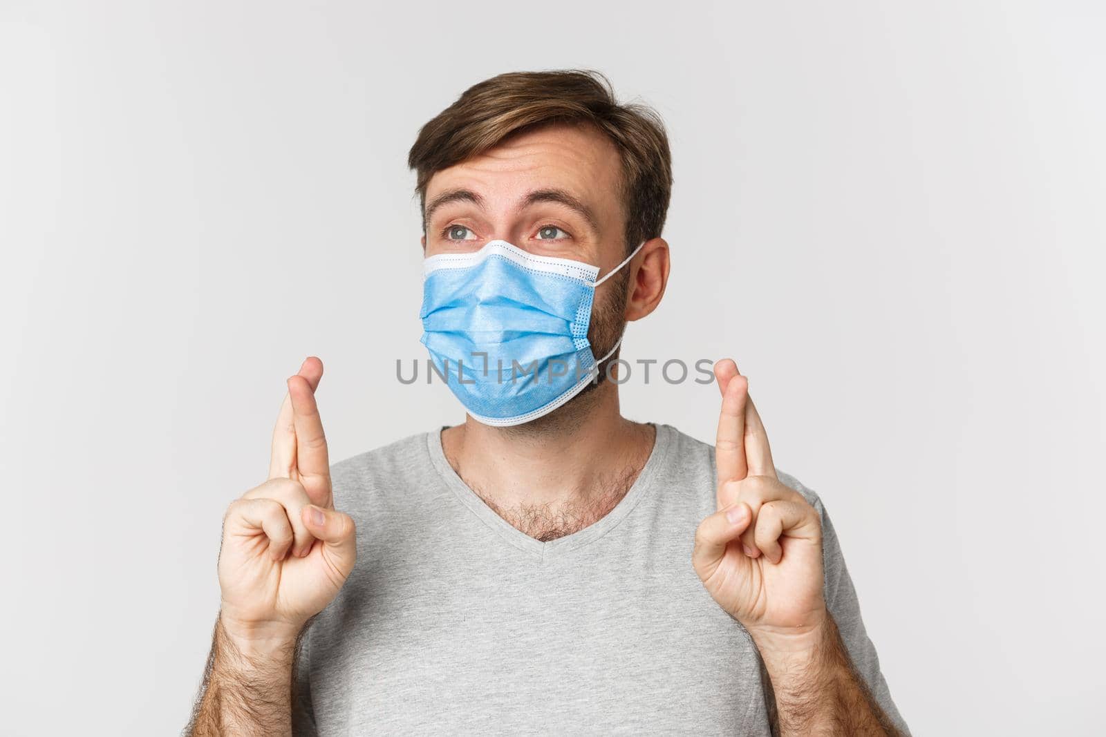 Concept of pandemic, covid-19 and social-distancing. Hopeful worried guy in medical mask, cross fingers for good luck and making wish, looking left, standing over white background.