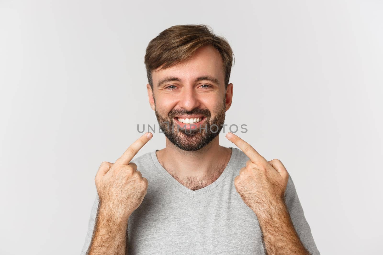 Close-up of handsome smiling man recommending dental clinic, pointing at his perfect white teeth, standing over white background by Benzoix