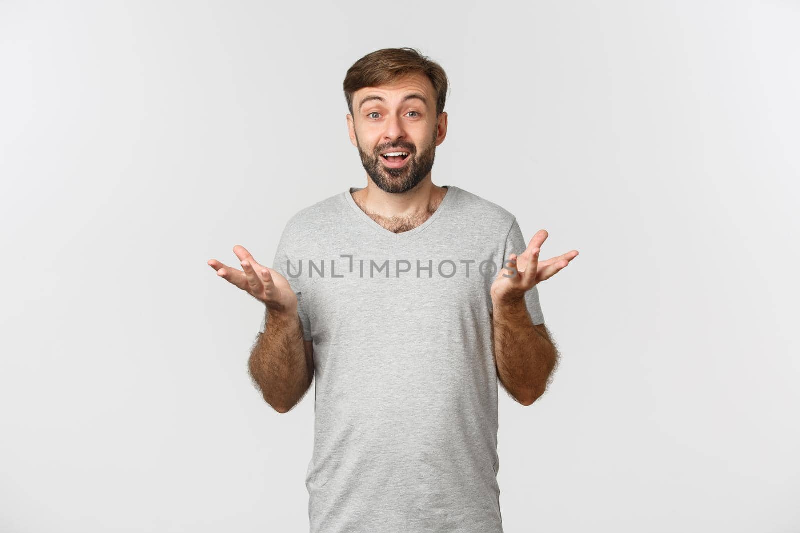 Portrait of handsome bearded male moden in grey t-shirt, raising hands up and looking amazed, congratulating you, standing over white background.
