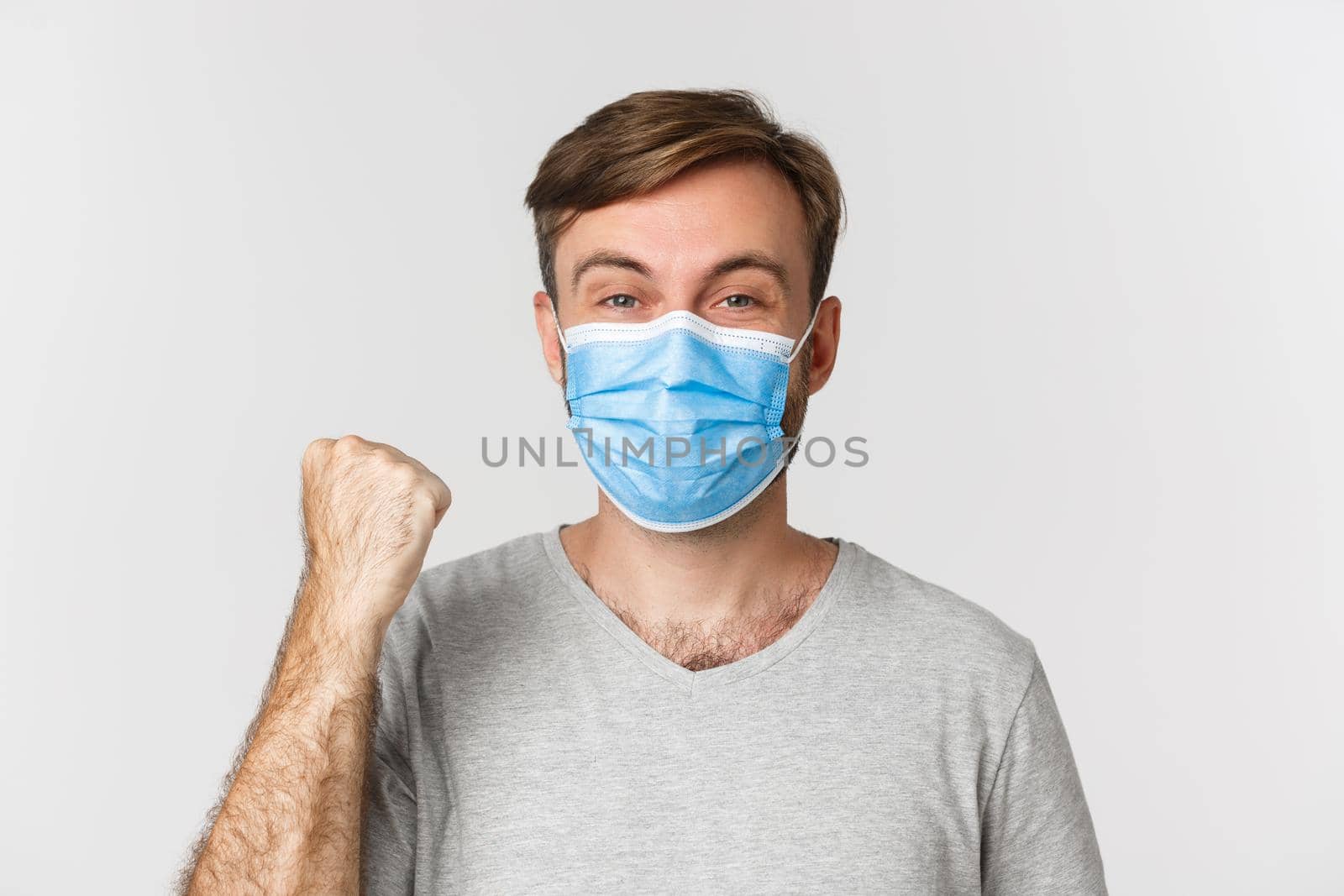 Concept of pandemic, coronavirus and social-distancing. Cheerful caucasian man in gray t-shirt and medical mask, rejoicing from win, standing over white background by Benzoix
