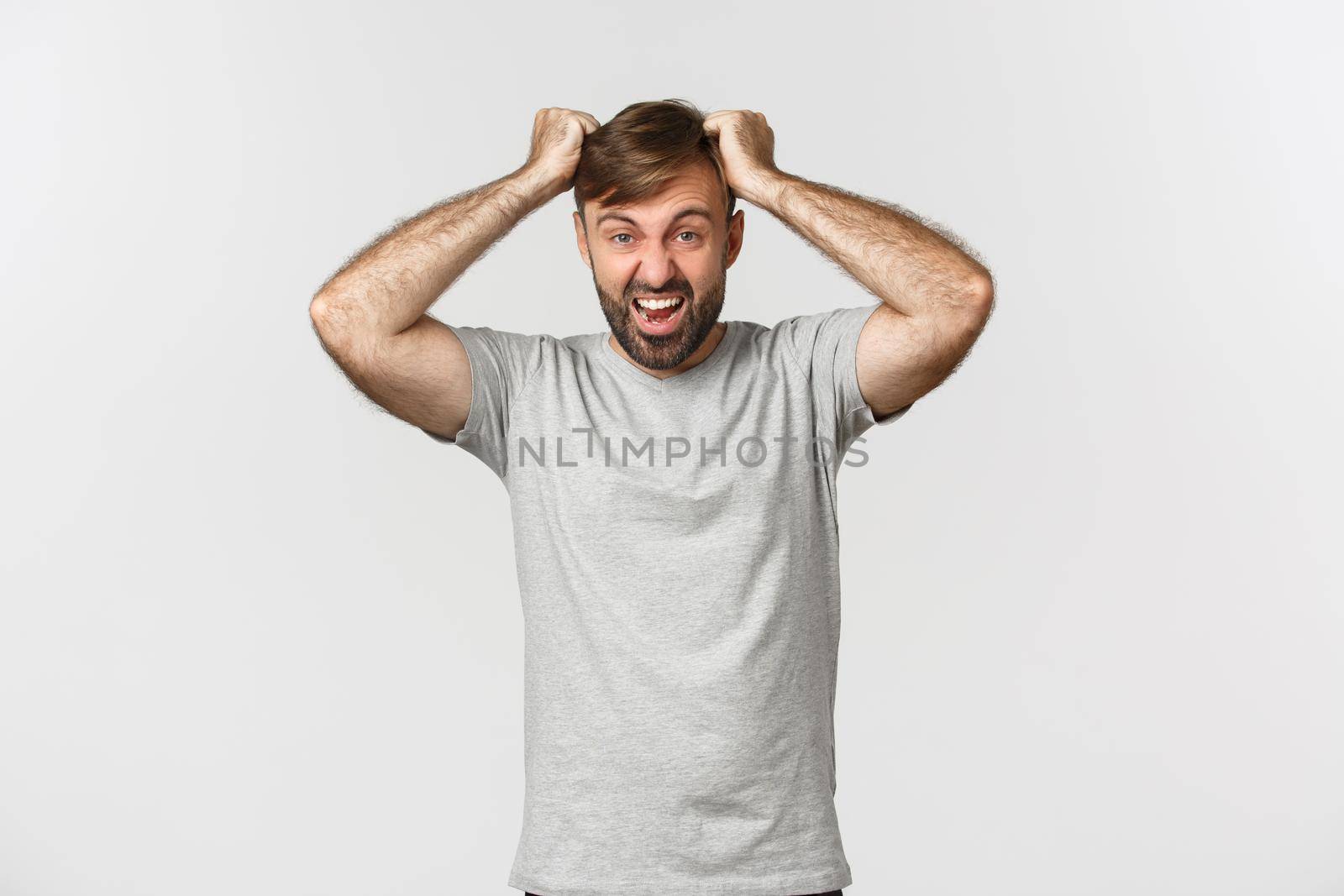 Portrait of distressed mad guy in gray t-shirt, ripping hair from anger and shouting, standing over white background.