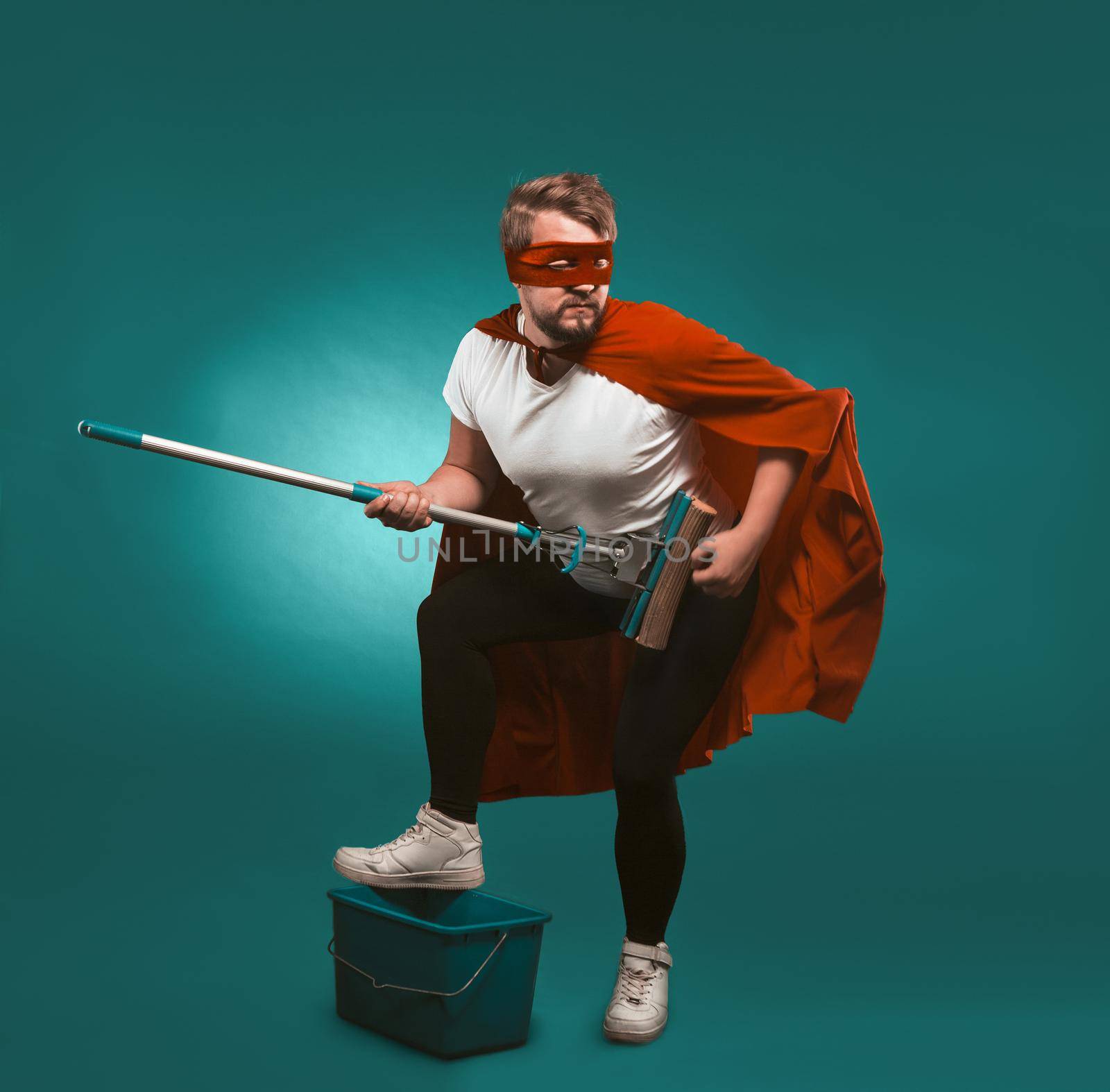 Superhero rock musician is cleaning. Man wearing a red cloak and mask holds a mop representing himself a musician putting his foot on a bucket. Cut out on biscay green background by LipikStockMedia