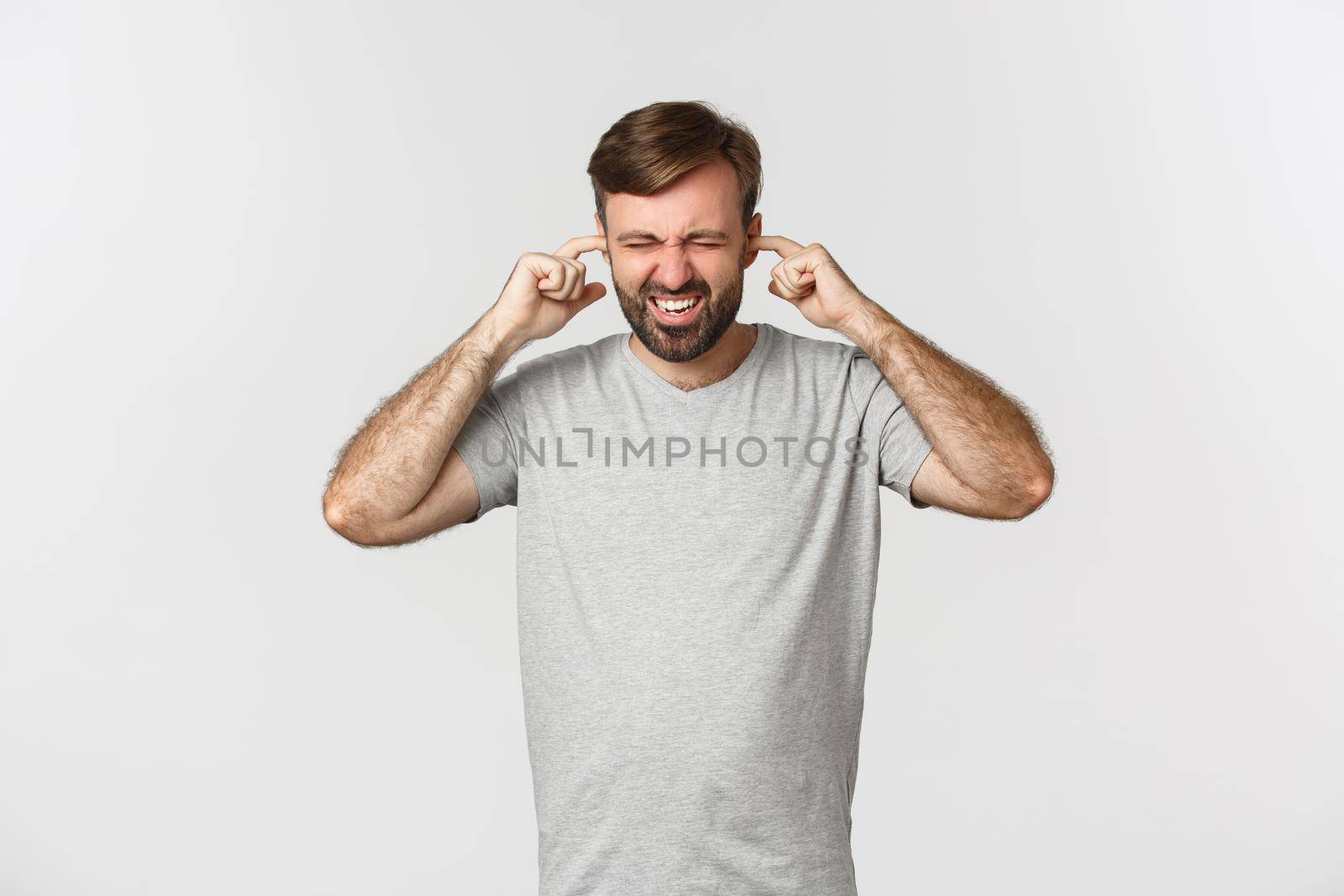 Portrait of annoyed guy in gray t-shirt, shutting ears and grimacing from loud noise, standing over white background by Benzoix