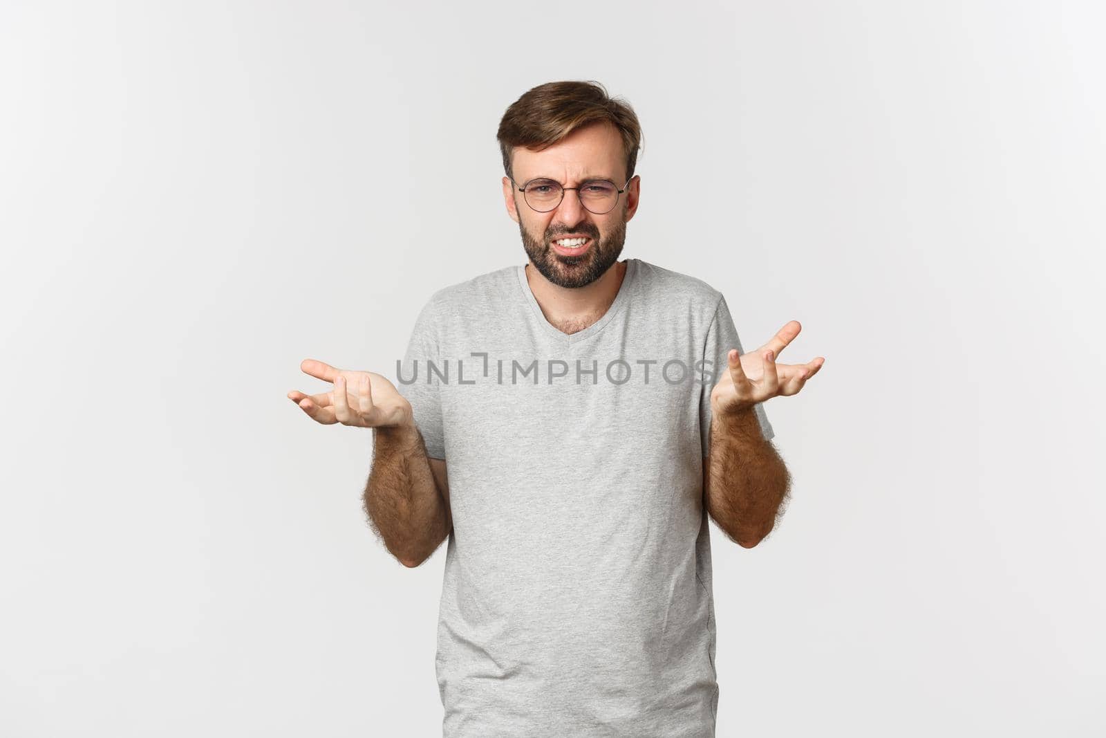 Image of frustrated young man in glasses and gray t-shirt, complaining, raising hands and looking pissed-off, standing over white background.