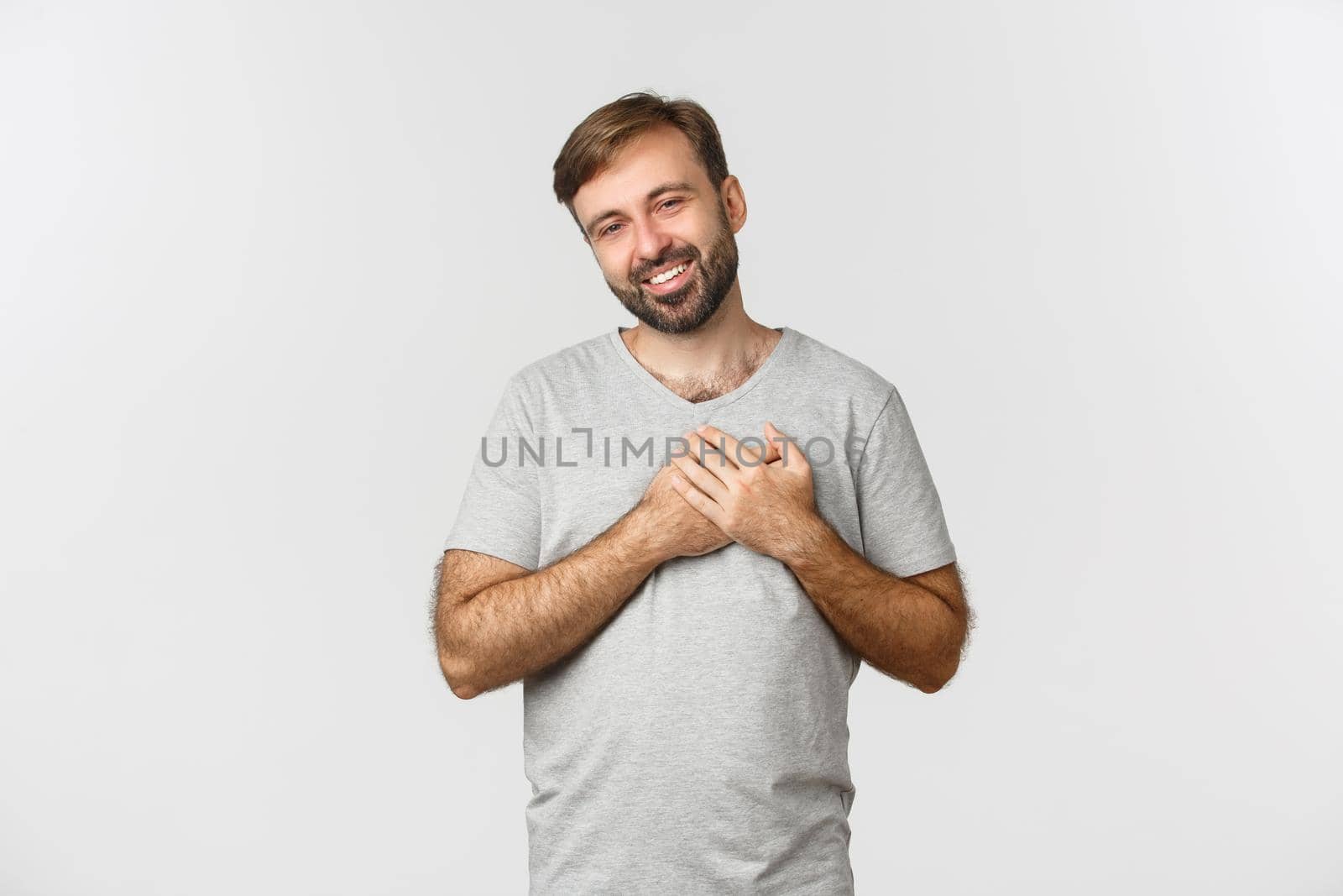 Image of handsome kind man with beard, wearing gray t-shirt, holding hands on heart and sighing, feeling thankful, express gratitude, standing over white background by Benzoix