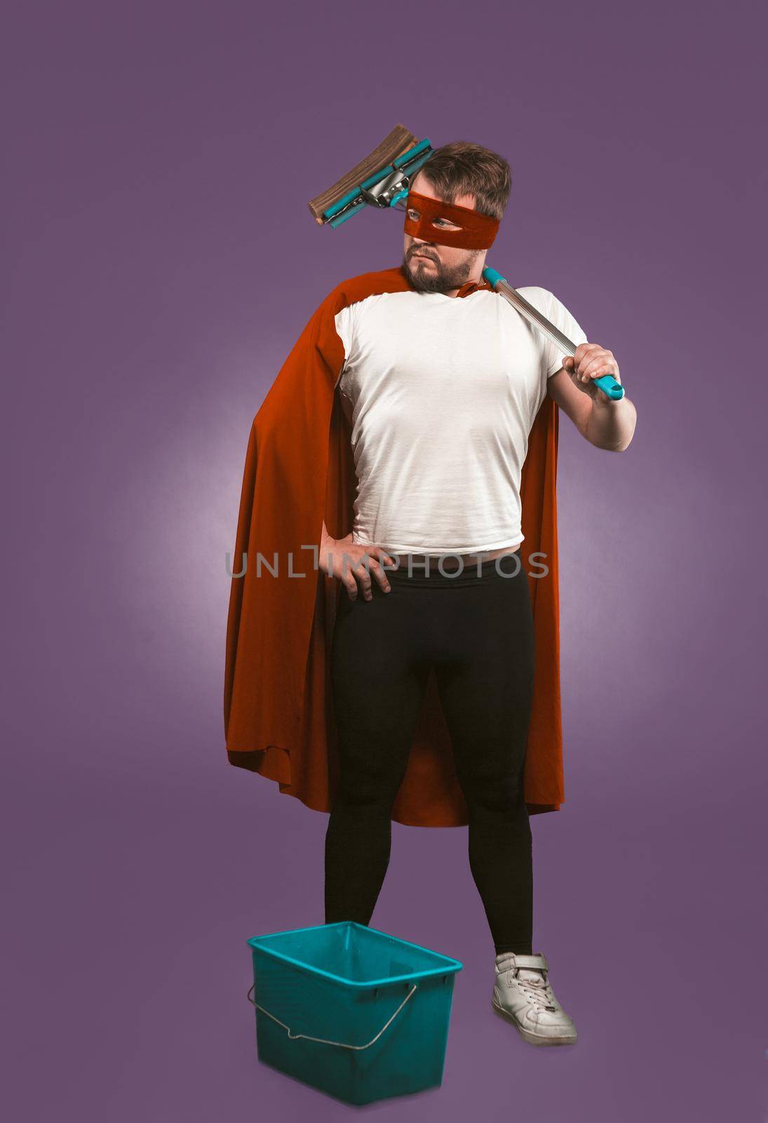 Super hero cleaner professional. Confidence man in red superhero costume with bucket and a mop stands on grape purple background. Super cleaning concept by LipikStockMedia