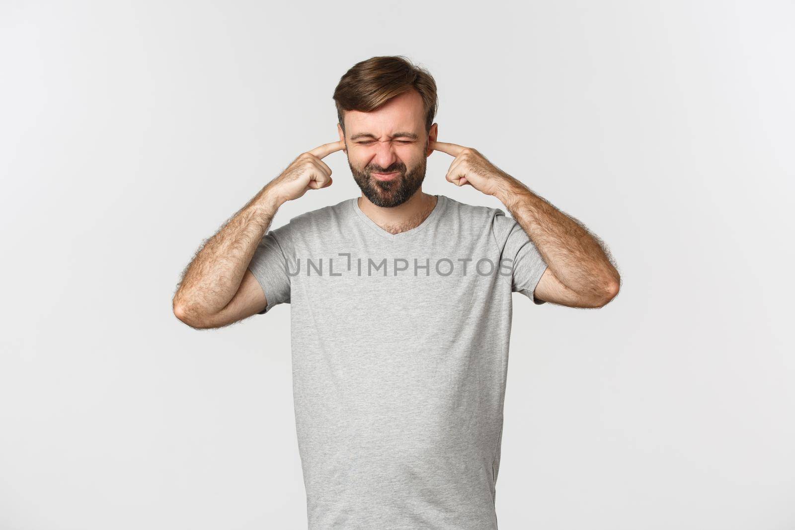 Portrait of annoyed guy in gray t-shirt, shutting ears and grimacing from loud noise, standing over white background.