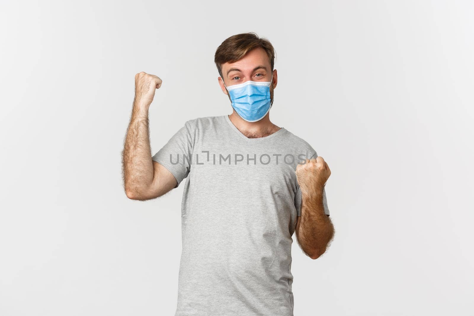 Concept of pandemic, coronavirus and social-distancing. Cheerful caucasian man in gray t-shirt and medical mask, winning something, rejoicing from win, standing over white background.
