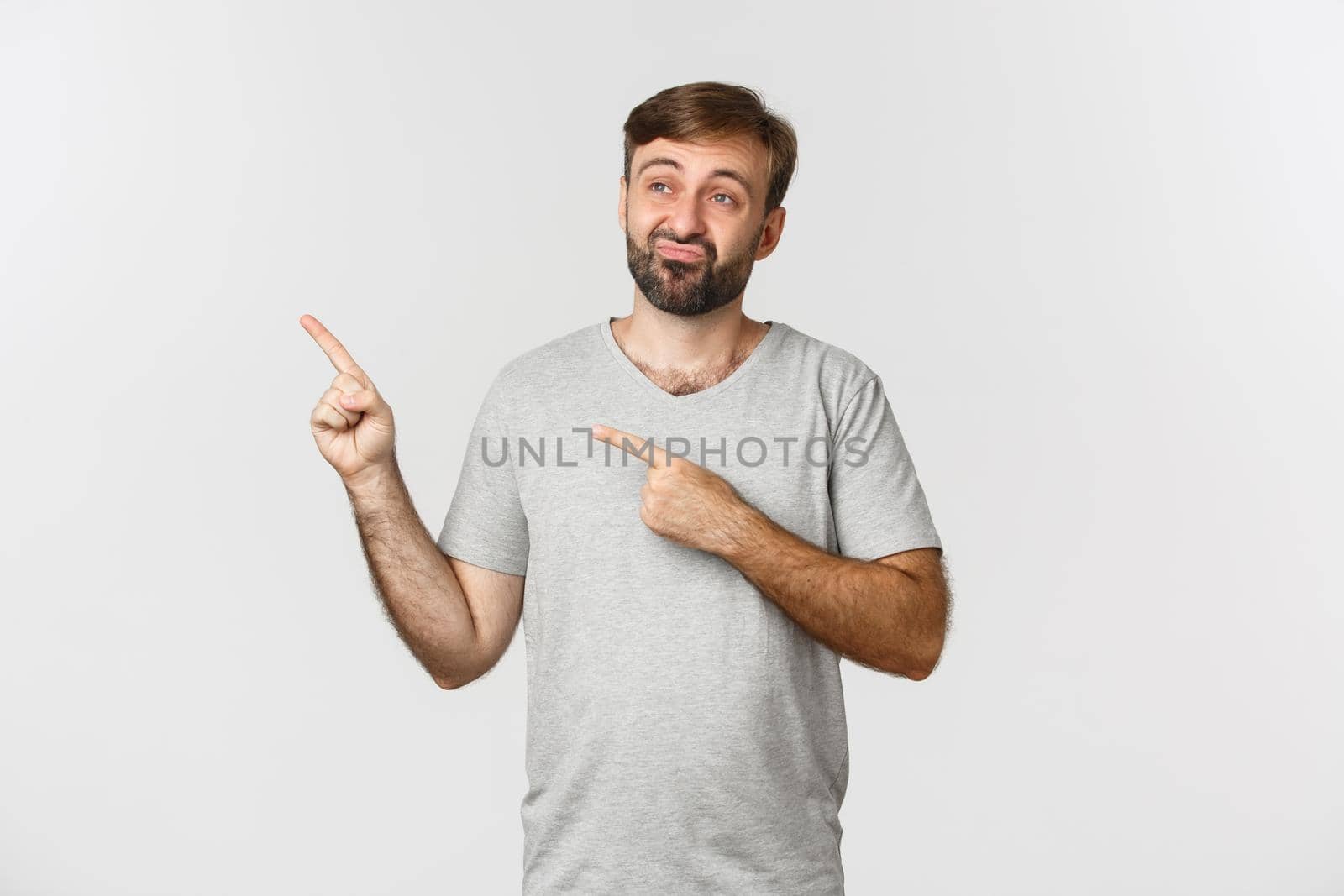 Image of skeptical and unamused bearded guy in gray t-shirt, pouting and pointing fingers at upper left corner, standing over white background by Benzoix