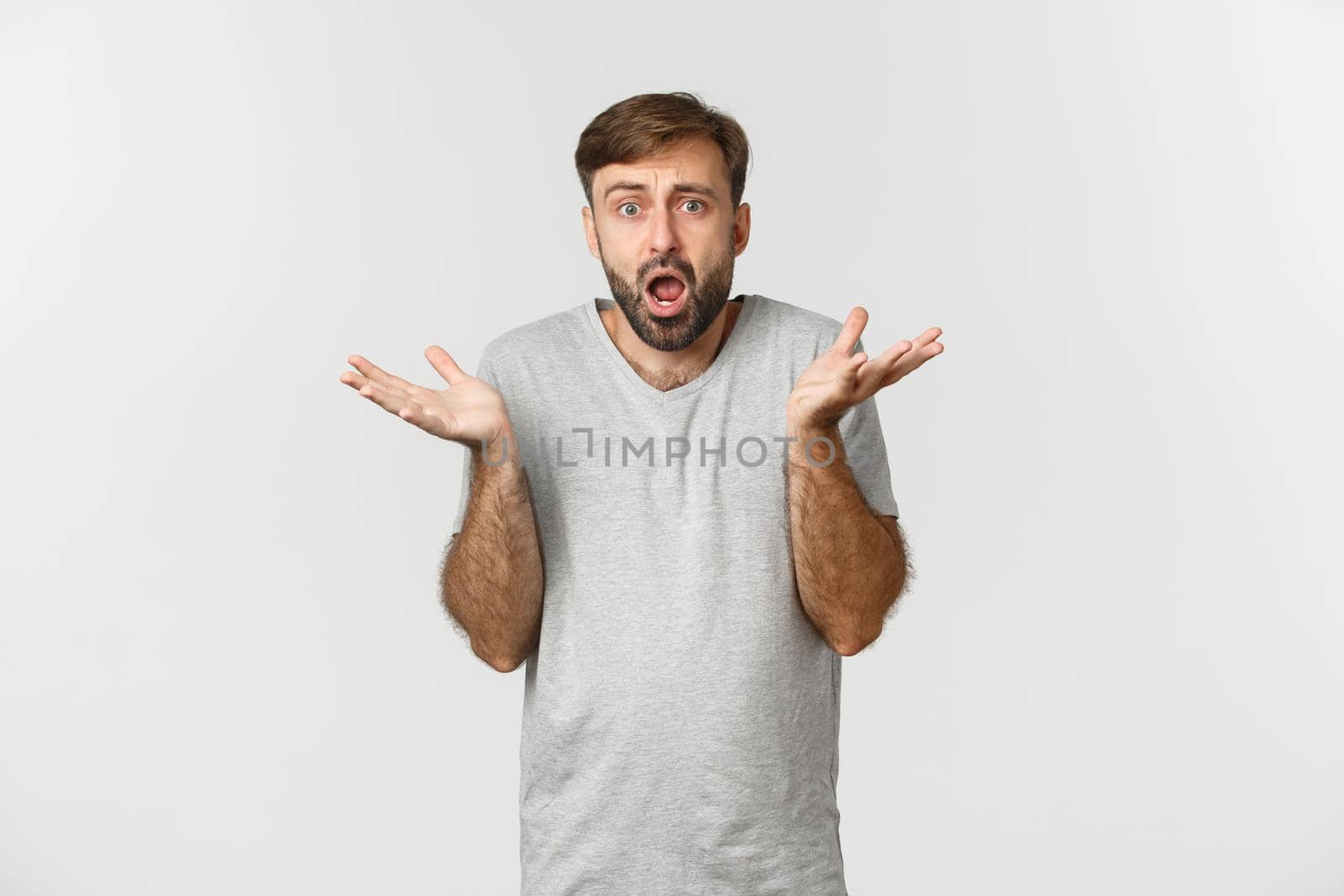 Portrait of confused and worried bearded guy, wearing casual t-shirt, looking at something complicated and shrugging, standing over white background.