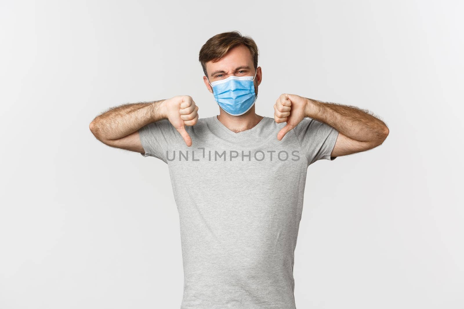 Concept of pandemic, covid-19 and social-distancing. Portrait of man hate wearing medical mask, complaining and showing thumbs-down, standing over white background by Benzoix