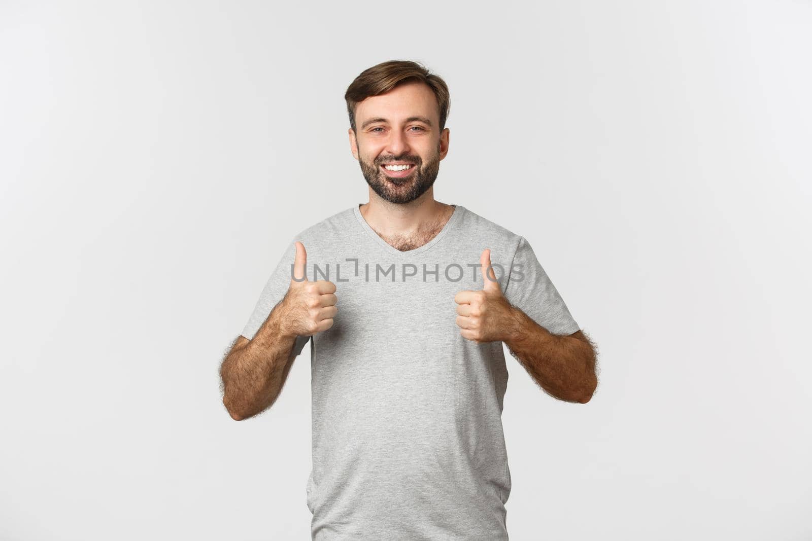 Image of handsome bearded man in gray t-shirt, showing thumbs-up in approval, like something good, standing over white background.
