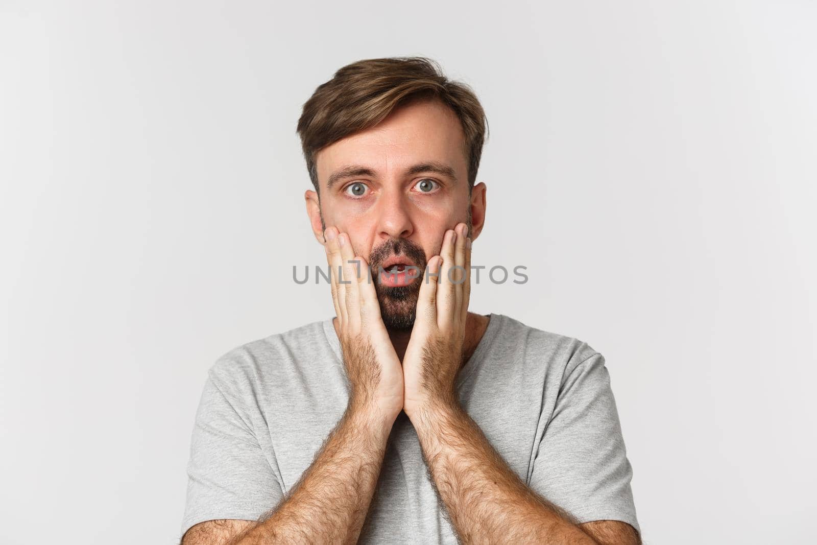 Close-up of shocked adult guy holding hands on face, gasping and looking at camera startled, standing over white background.