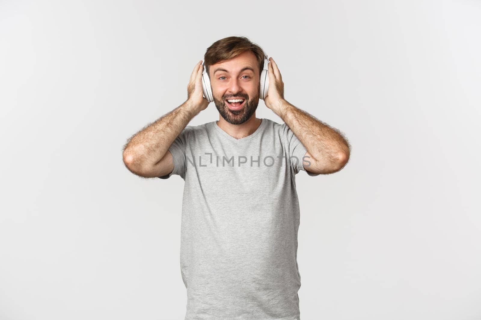 Image of handsome man in casual outfit, listening music in wireless headphones, smiling pleased, standing over white background.