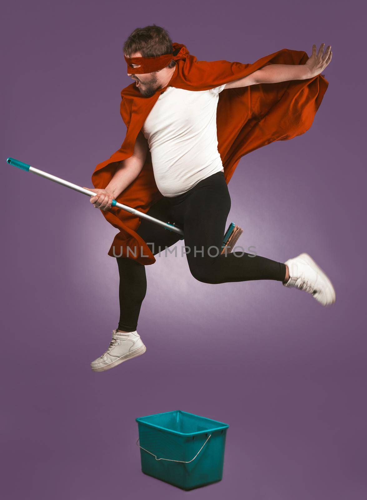 Super hero flying on mop. Funny man have fun with a bucket and a mop on grape purple background. Funny cleaning concept.