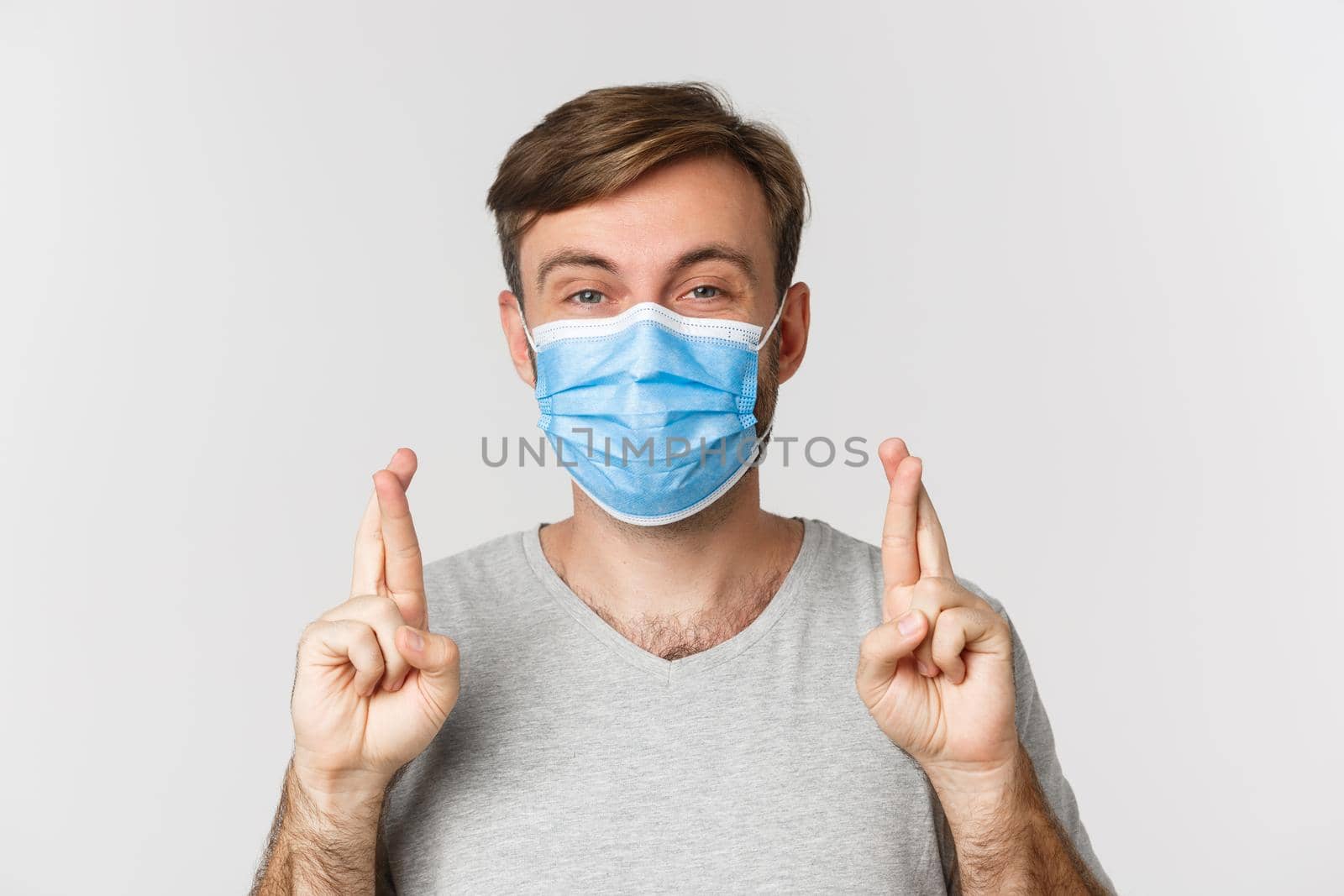 Concept of pandemic, covid-19 and social-distancing. Hopeful worried guy in medical mask, cross fingers for good luck and making wish, standing over white background by Benzoix