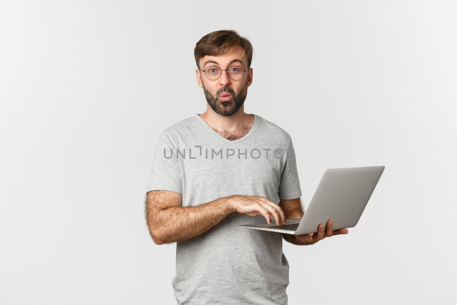 Image of surprised male model in glasses and gray t-shirt, looking amazed, using laptop, standing over white background.