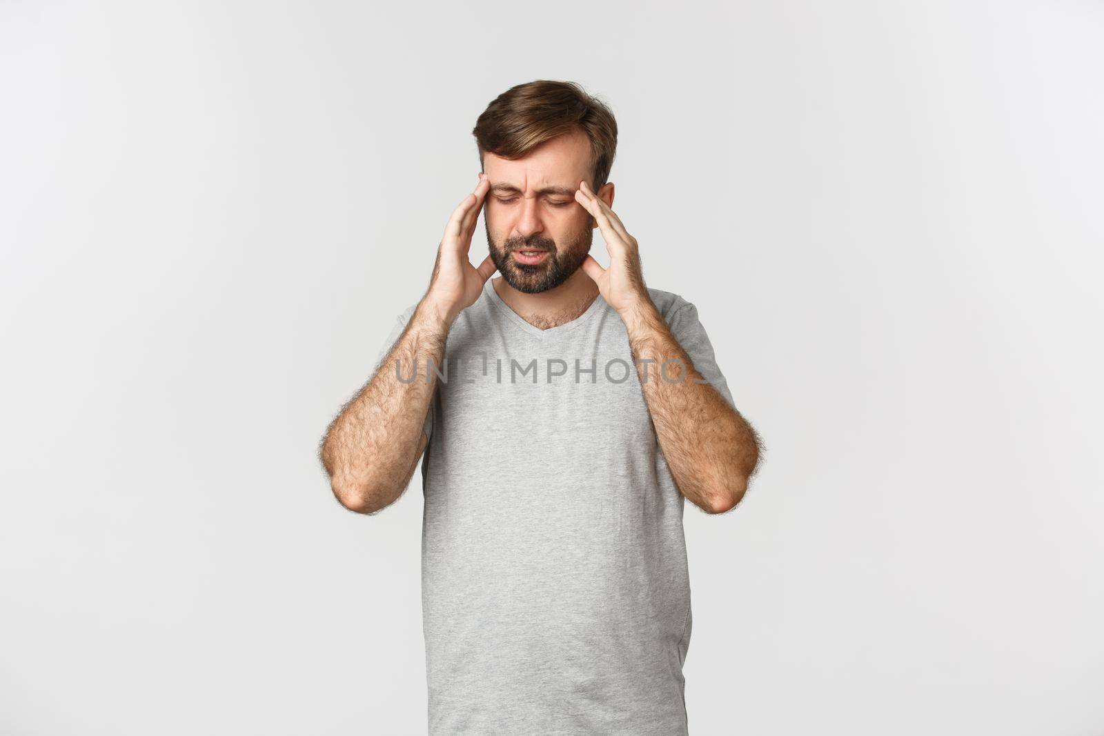 Man having a severe headache, grimacing and touching head, feeling dizzy or sick, standing in gray t-shirt over white background.