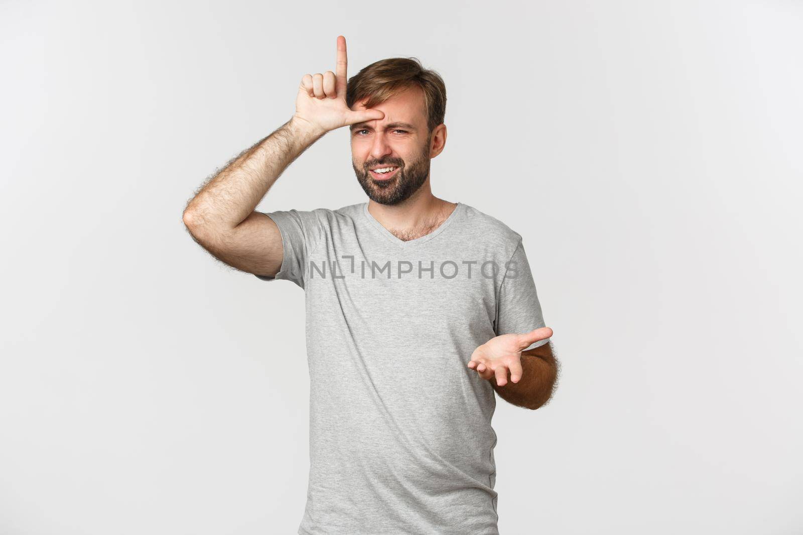 Portrait of arrogant man in gray t-shirt, mocking person who lost, showing loser sign on forehead and looking with dismay, standing over white background.