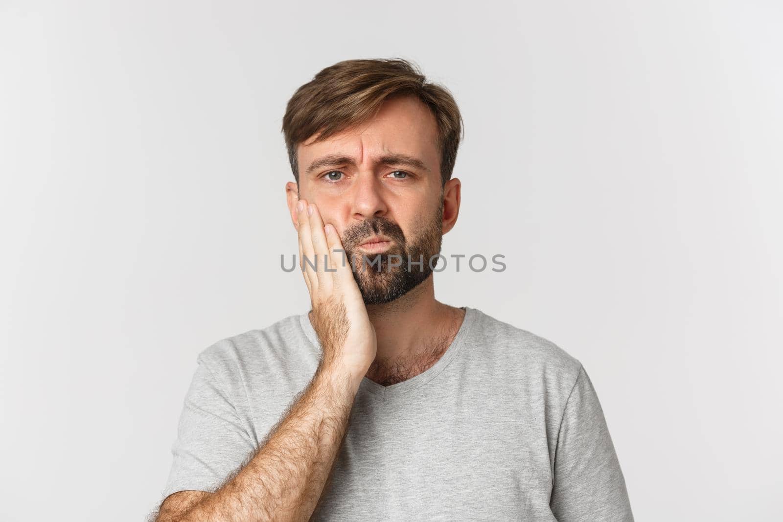 Close-up of gloomy bearded man, touching cheek and complaining on toothache, need see dentist, standing over white background.