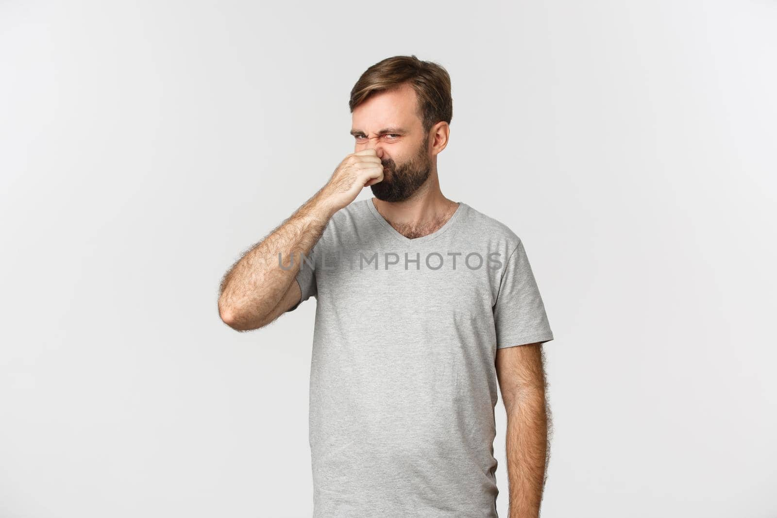 Portrait of disgusted caucasian man in gray t-shirt, shut his nose from awful smell, standing over white background.