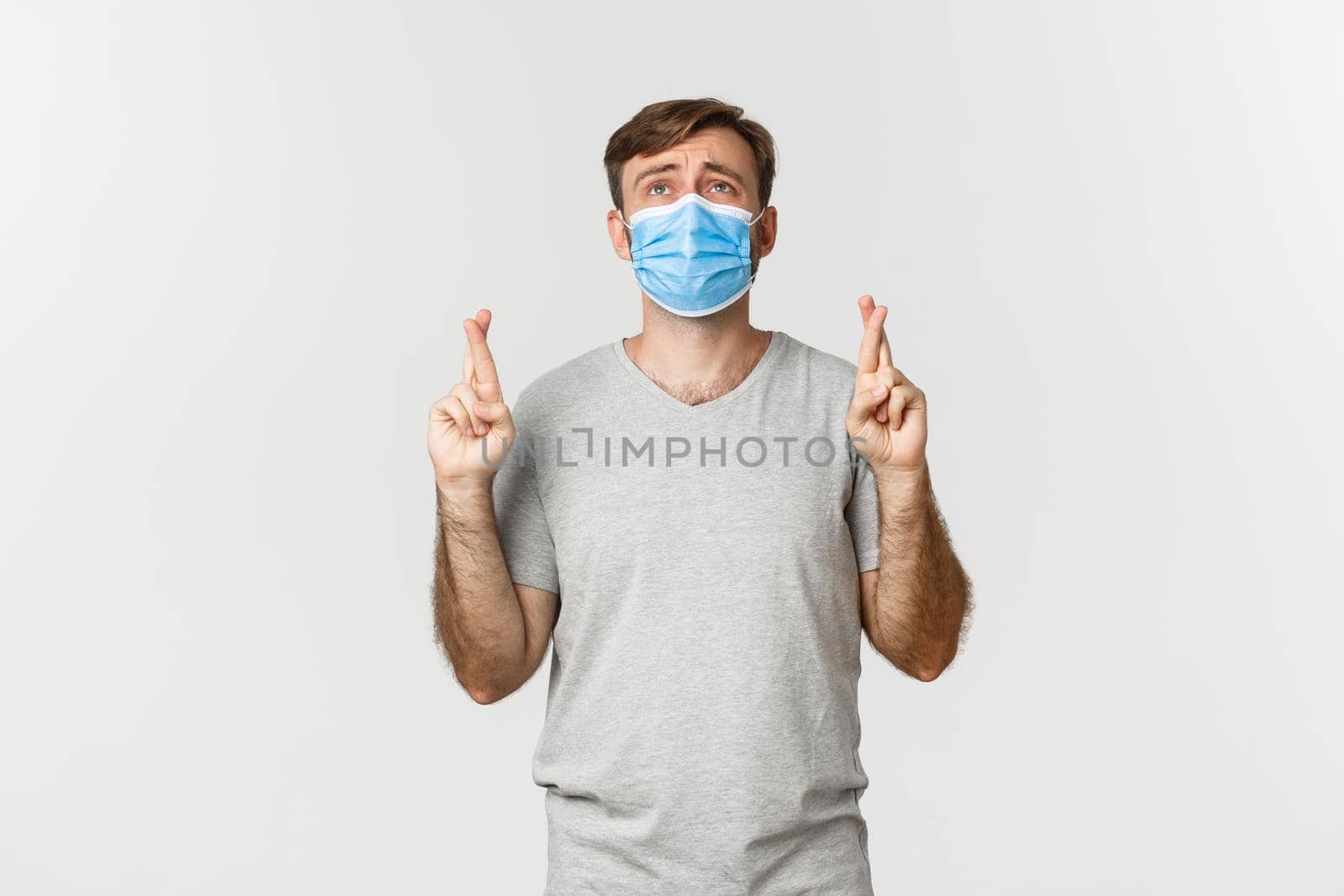 Concept of pandemic, covid-19 and social-distancing. Hopeful worried guy in medical mask, cross fingers for good luck and begging god, looking up desperate, standing over white background by Benzoix