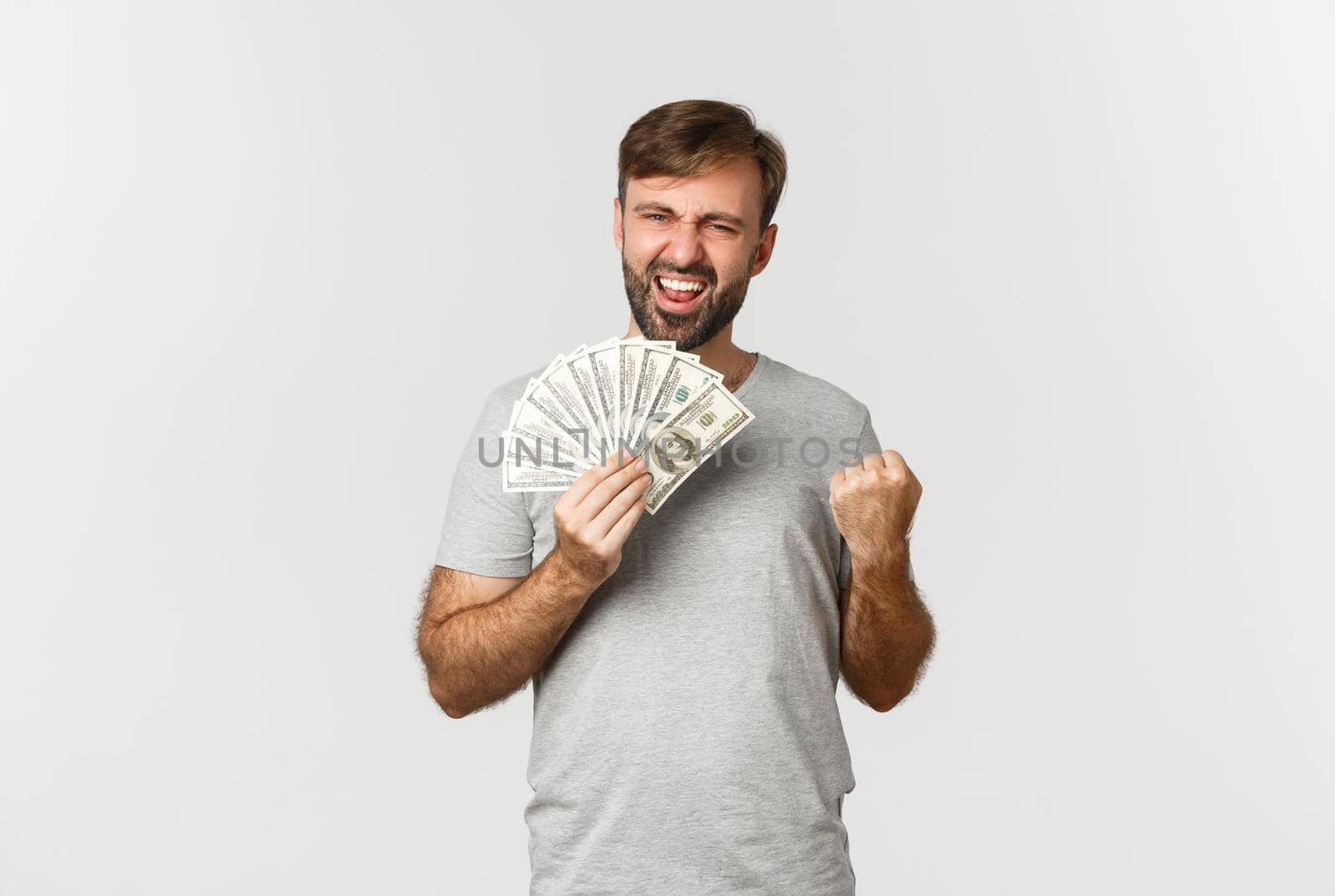 Pleased smiling man with beard, wearing gray t-shirt, rejoicing and winning cash, holding money and celebrating, standing over white background.