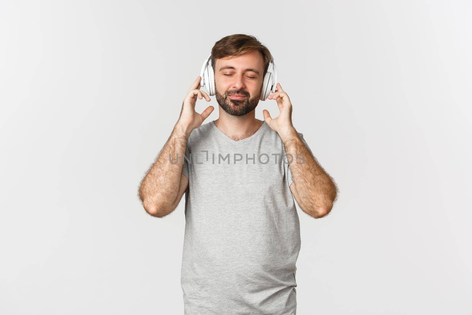 Image of handsome bearded man in gray t-shirt, close eyes and smiling with satisfaction of music, listening to song in headphones, standing over white background.