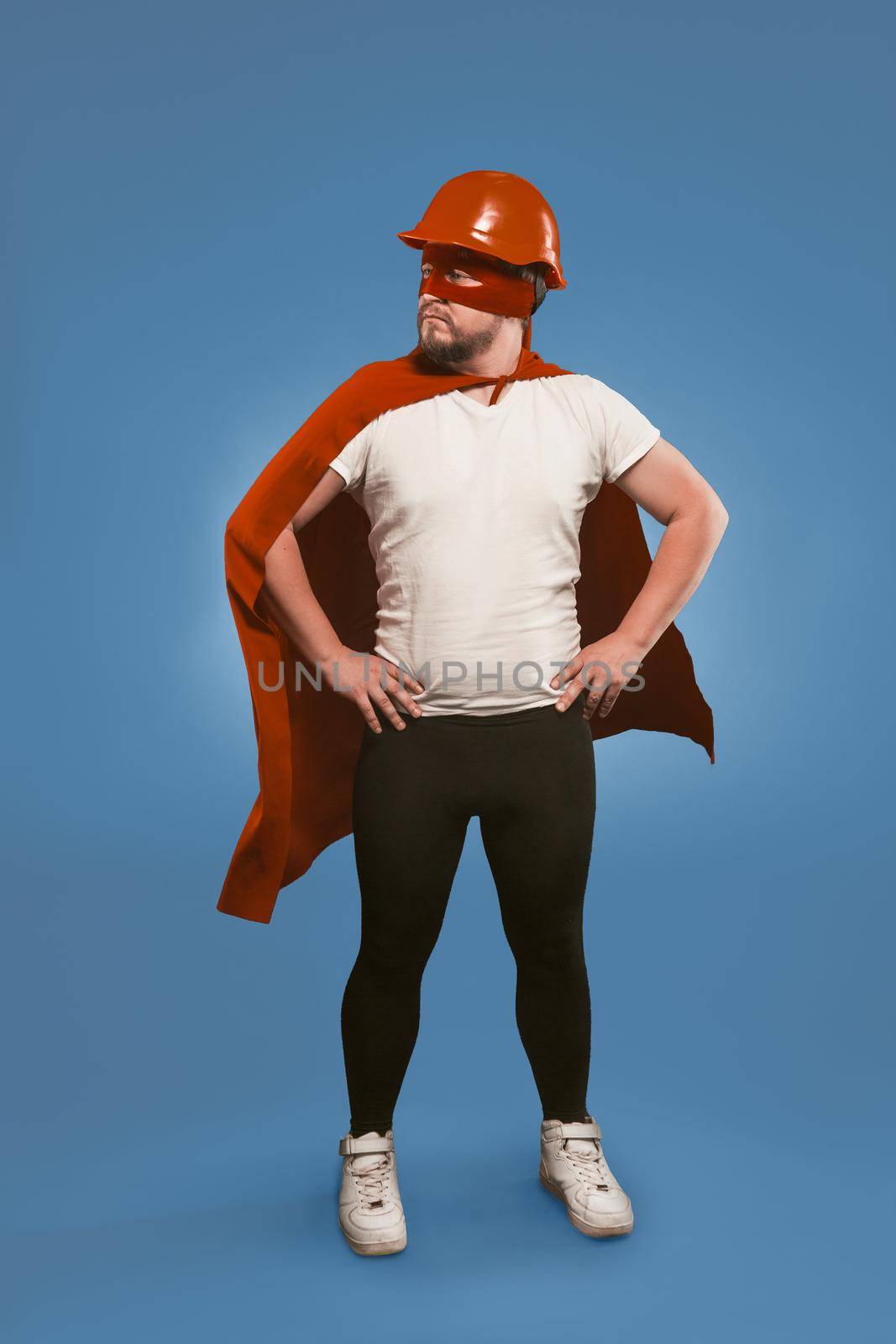 Super hero engineer. Man wearing red helmet, mask and cape standing with his hands on hips. Isolated on faded denim blue background by LipikStockMedia