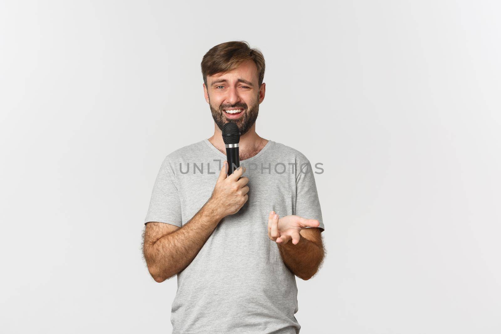 Image of guy with beard, wearing gray t-shirt, making speech with microphone and looking confused, standing over white background.