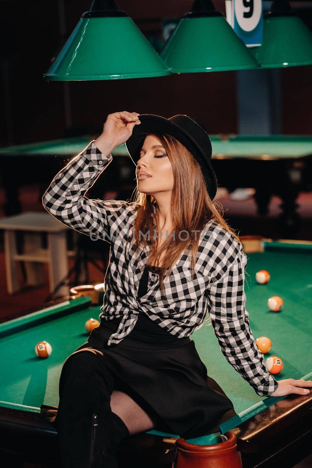 A girl in a hat in a billiard club sits on a billiard table.Playing pool by Lobachad
