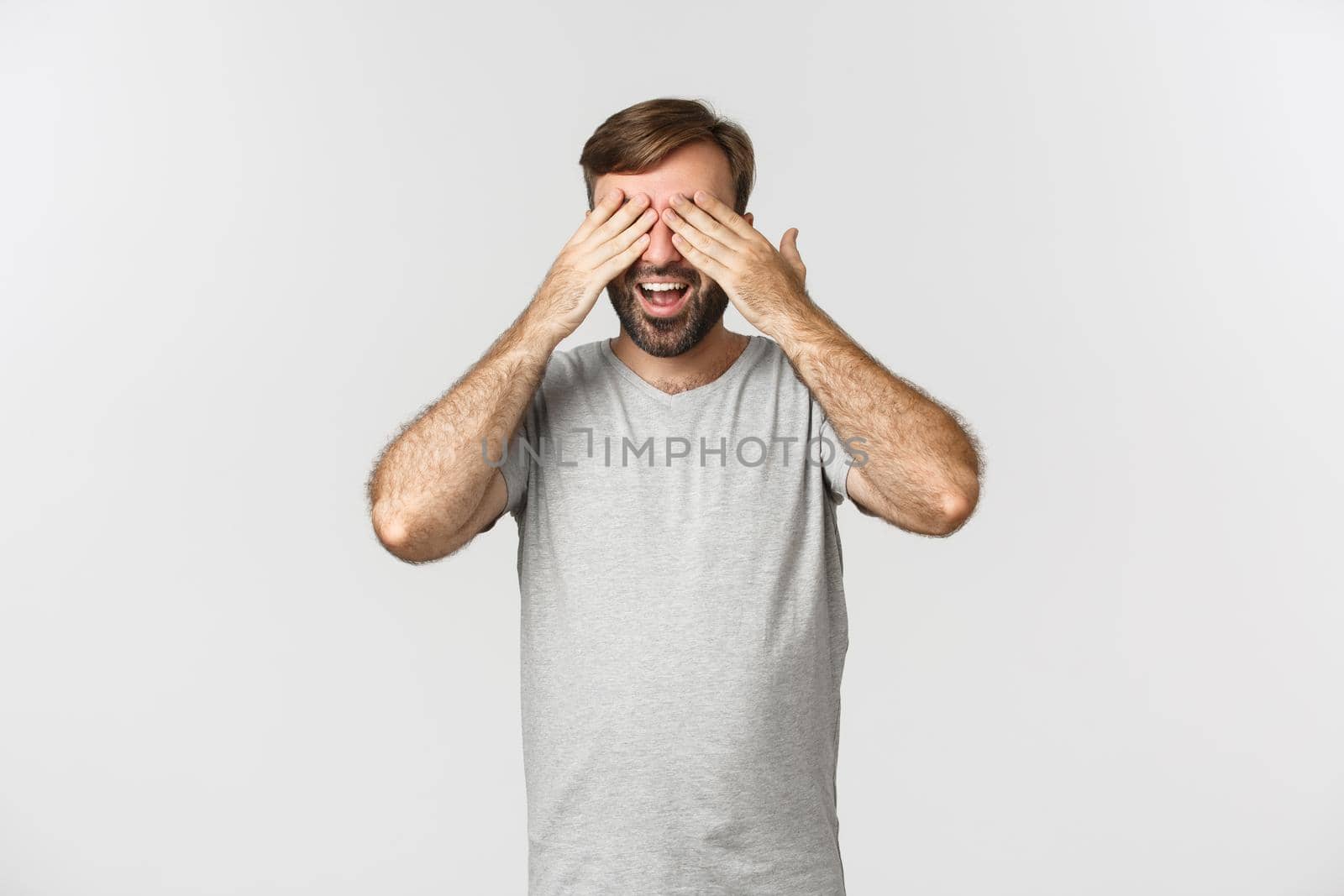 Image of happy smiling guy with beard, close eyes and waiting for surprise, anticipating something, standing over white background.
