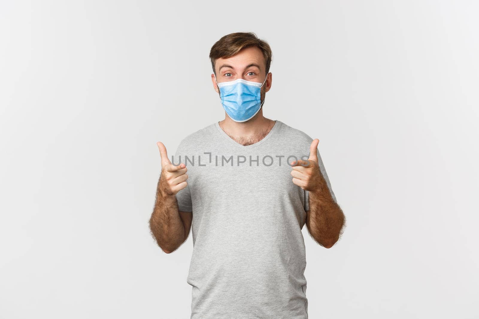 Concept of pandemic, coronavirus and social-distancing. Excited caucasian man in medical mask and gray t-shirt, pointing fingers at camera and praising you, standing over white background.