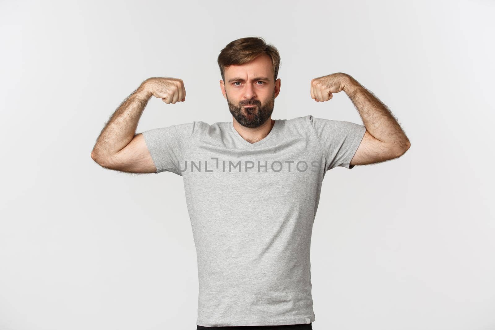 Portrait of confident bearded man flexing biceps, showing his muscles after workout, standing over white background by Benzoix